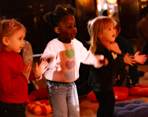 Es sind kleine Kinder zu sehen, die aufmerksam zuhören, tanzen und in die Hände klatschen. Fotocredit: Christoph Brunkow